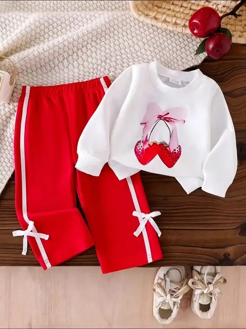 Este conjunto de ropa para niñas incluye una sudadera blanca con un estampado de lazo y unos pantalones rojos con franjas blancas laterales.
