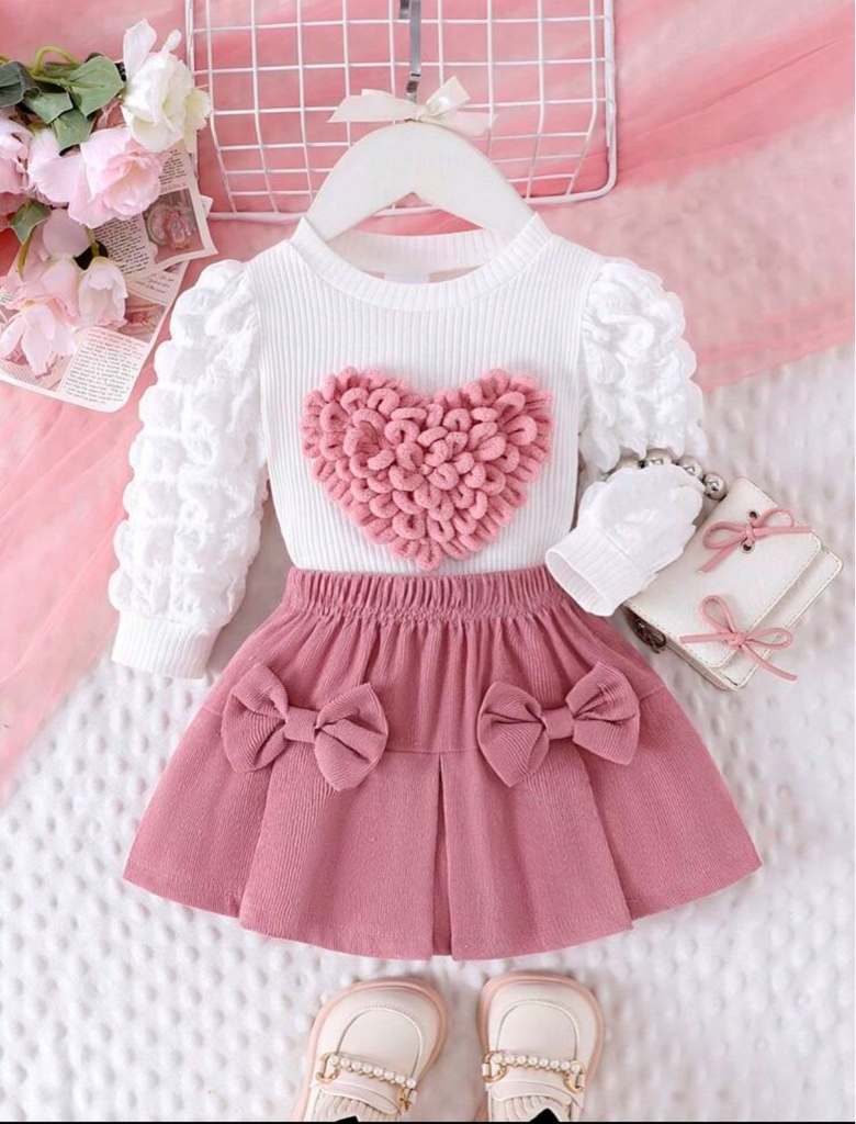 Este conjunto de ropa para niña de 2 piezas incluye una blusa blanca con mangas abullonadas y una falda rosa acanalada con lazos.