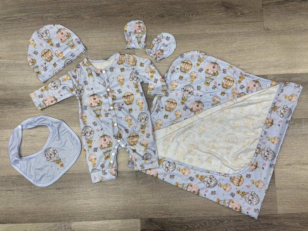 Este conjunto de mameluco para bebé presenta un estampado caprichoso de globos aerostáticos y elefantes en tonos pastel sobre una base azul claro.