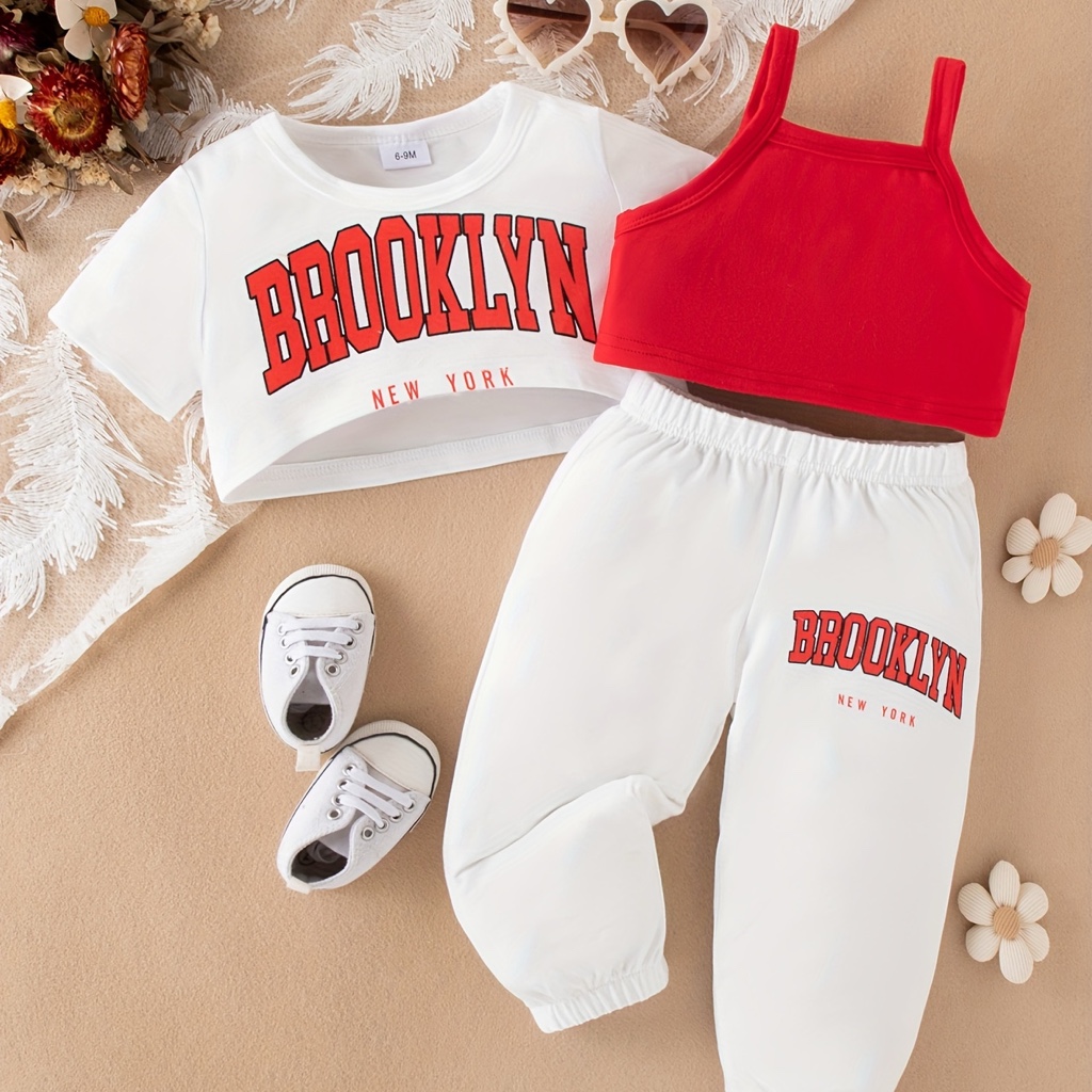 Este conjunto de ropa para niña cuenta con una camiseta blanca corta con la palabra "BROOKLYN" en rojo y pantalones blancos a juego.