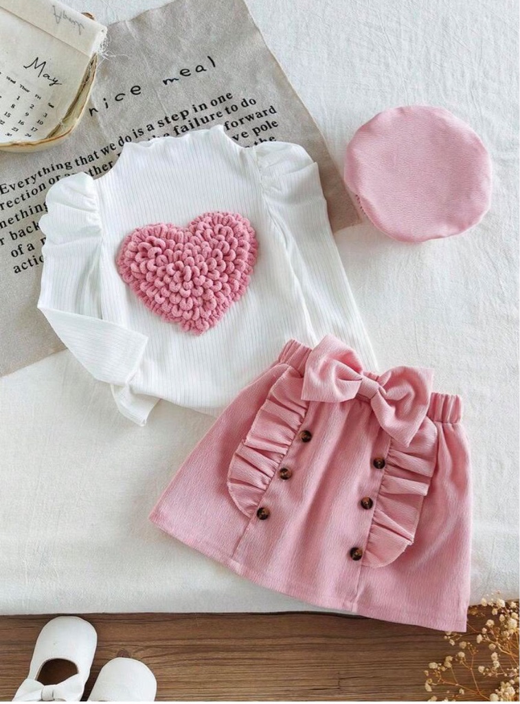 Este conjunto de tres piezas para niña de incluye una blusa blanca de cuello alto con un aplique de corazón texturizado en 3D